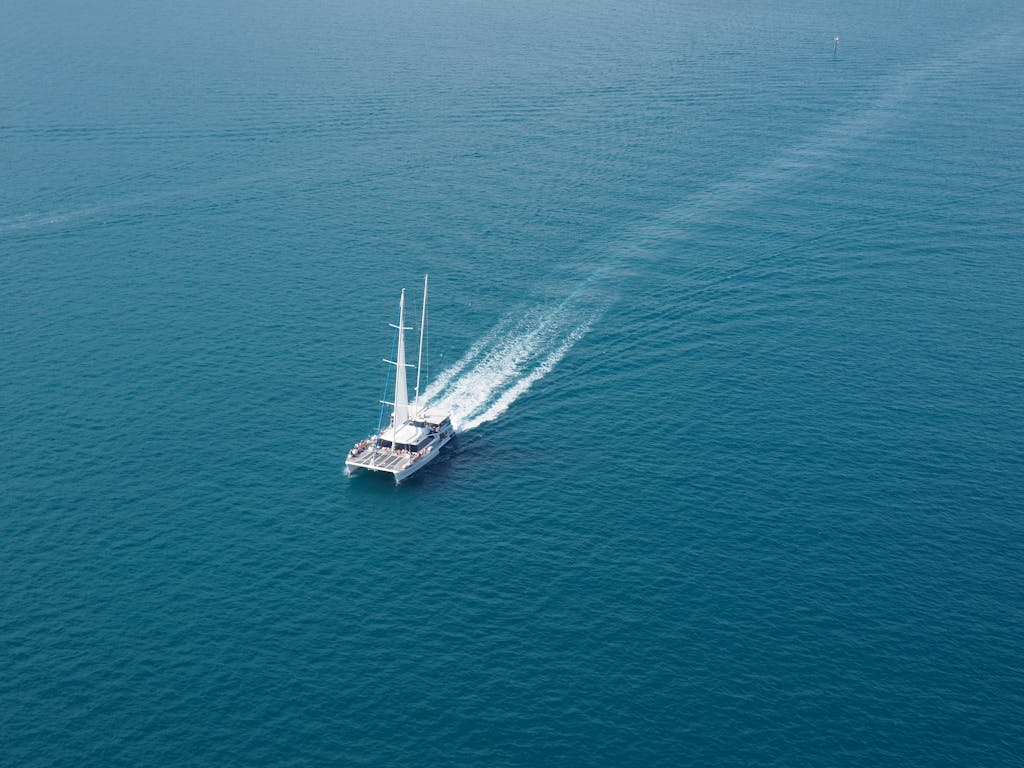 A sailboat cruises through clear blue ocean waters, captured from an aerial perspective.